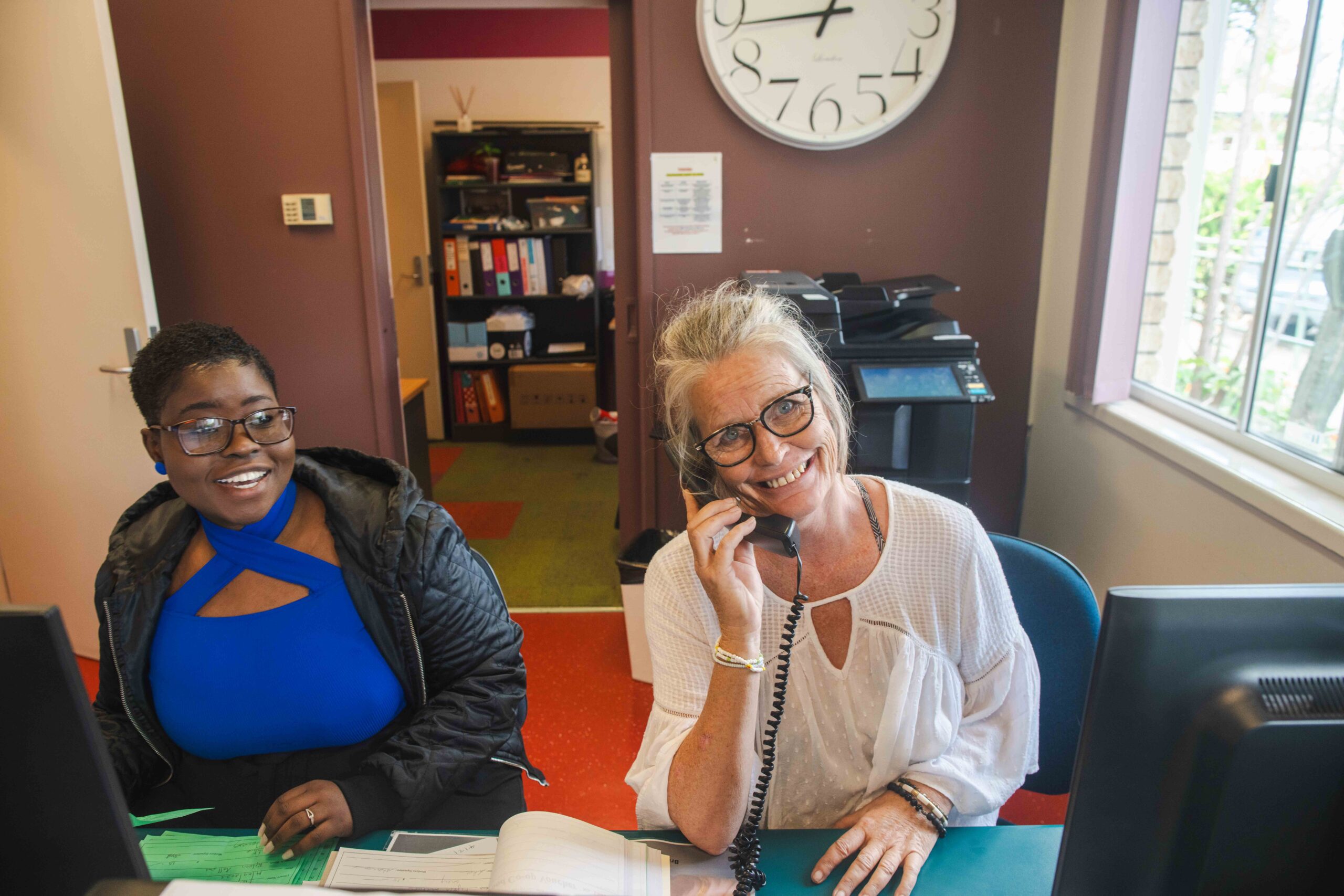 Two women working in an office setting. One woman, with short black hair and glasses, is wearing a blue top and a black jacket, smiling at the camera. The other woman, with long gray hair and glasses, is speaking on the phone while wearing a white blouse, also smiling. A clock and office equipment are visible in the background.