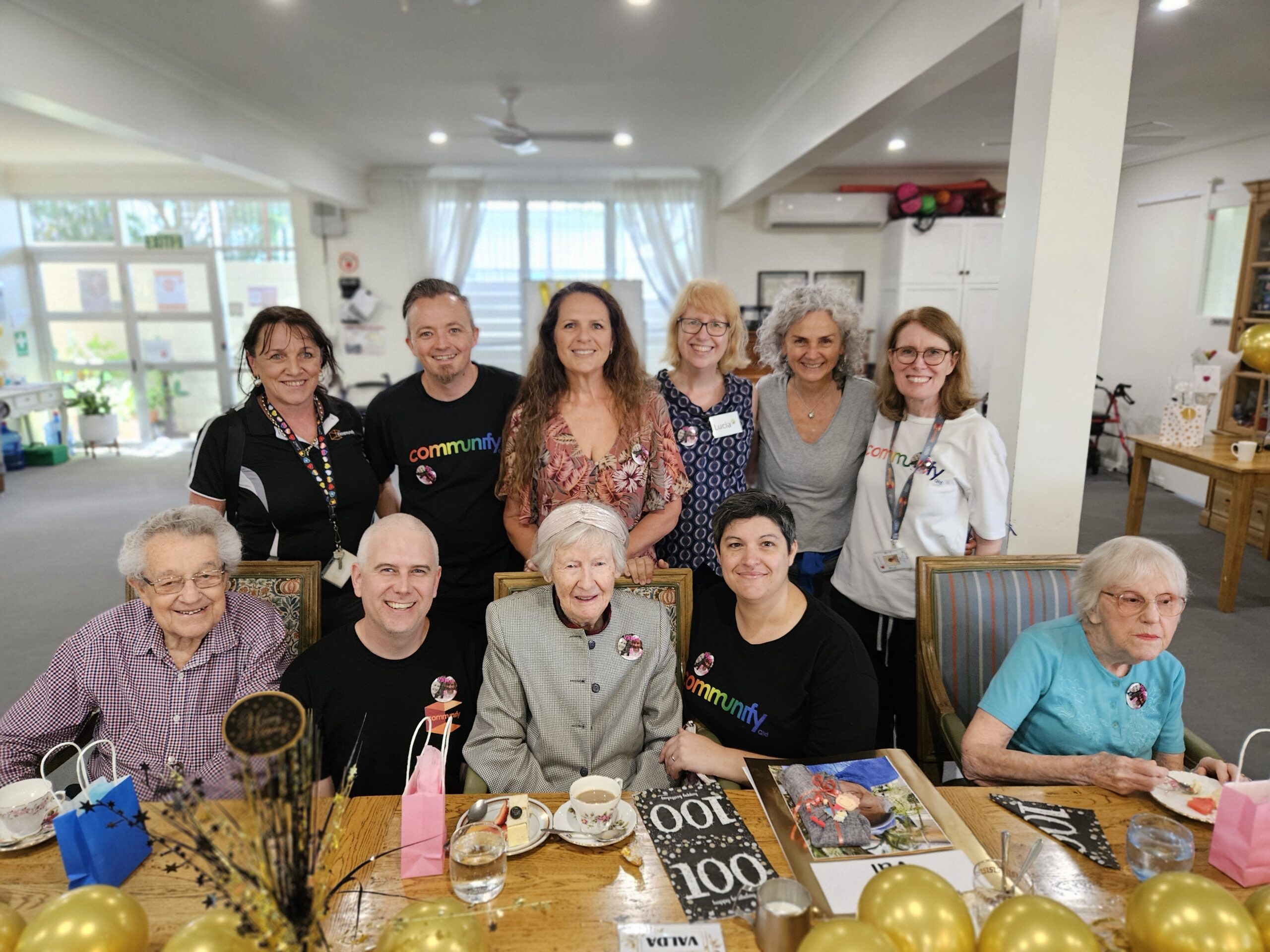 Group photo of a celebration with a diverse group of people. In the front, an elderly woman wearing a grey sweater and smiling, surrounded by younger individuals. The back row includes women in various outfits, including black and colorful shirts, showcasing a mix of ethnicities and cheerful expressions. Decorations and magazines are visible on the table, hinting at a festive occasion.