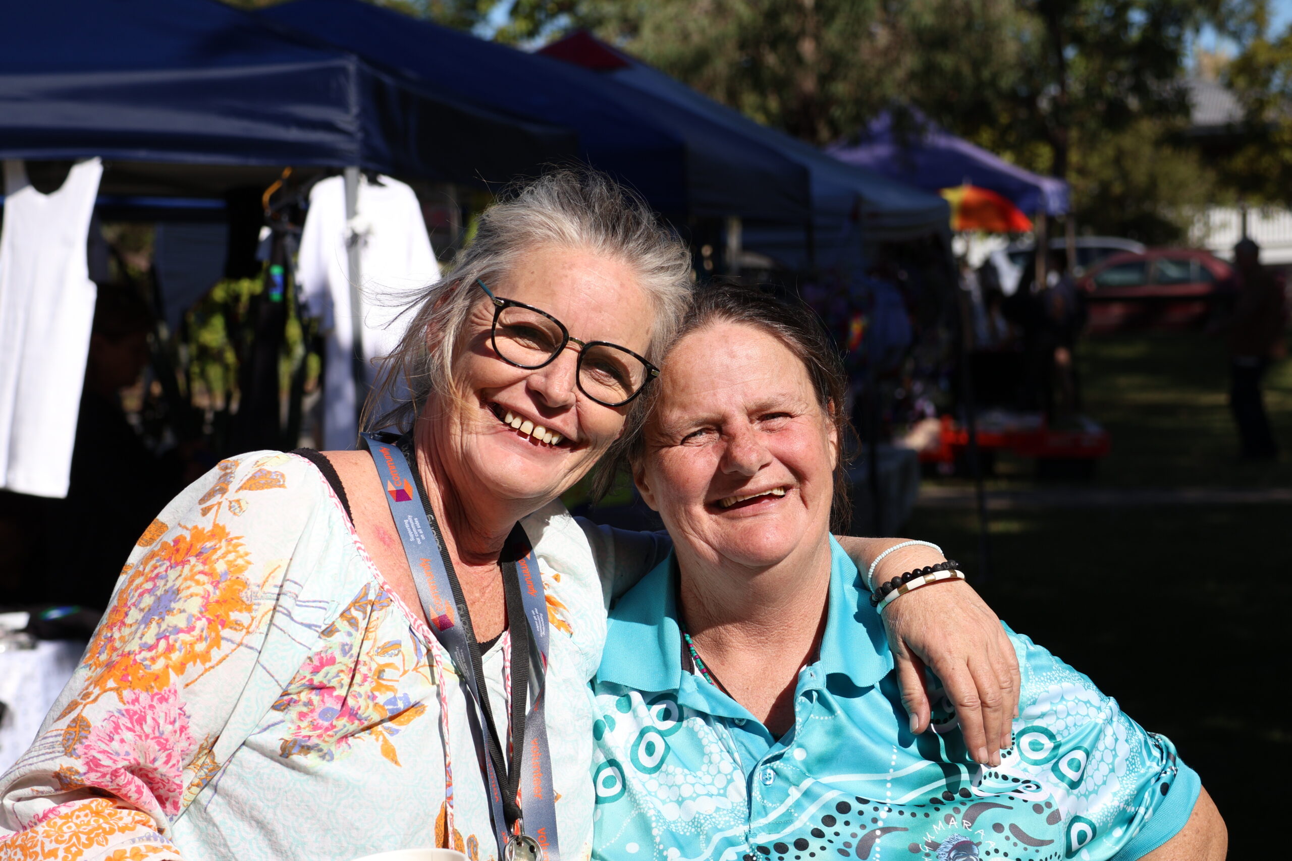Two women smiling and posing for the camera. The woman on the left has gray hair and wears glasses, dressed in a colorful patterned shirt. The woman on the right has dark hair and is wearing a turquoise shirt. Both appear happy and friendly.