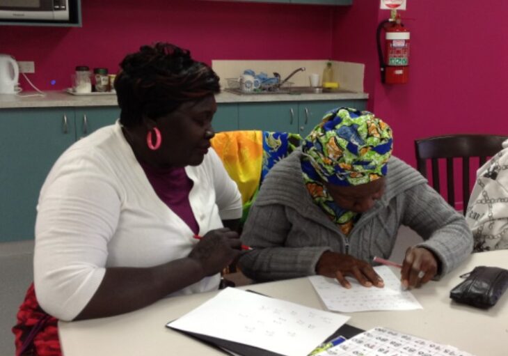 Two Black women are engaged in a conversation at a table. One woman, wearing a white shirt and pink earrings, is looking attentively at the other, who is wearing a colorful headscarf and a gray sweater. The second woman is focused on writing on a piece of paper.