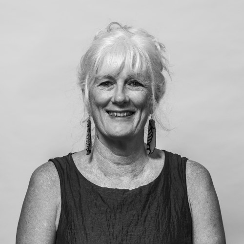 Smiling older woman with white hair styled in loose waves, wearing a dark-colored sleeveless dress. She has large earrings and a warm, friendly expression.