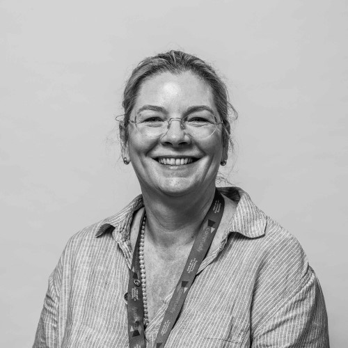 Smiling middle-aged woman with light skin, wearing a gray button-up shirt and a colorful lanyard, against a plain background.