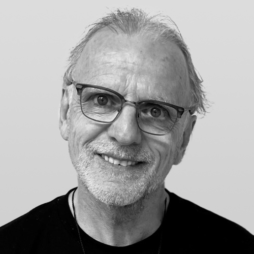 Smiling older Caucasian man with gray hair and glasses, wearing a black shirt. He has a friendly expression, showcasing warmth and approachability.