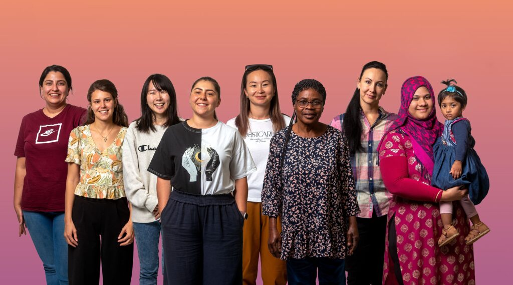 Group of seven women standing together, showcasing diverse ethnicities. They are dressed in a variety of clothing styles: a white t-shirt, a black graphic tee, a patterned blouse, and a pink plaid shirt. Their expressions are friendly and approachable, reflecting a sense of camaraderie.