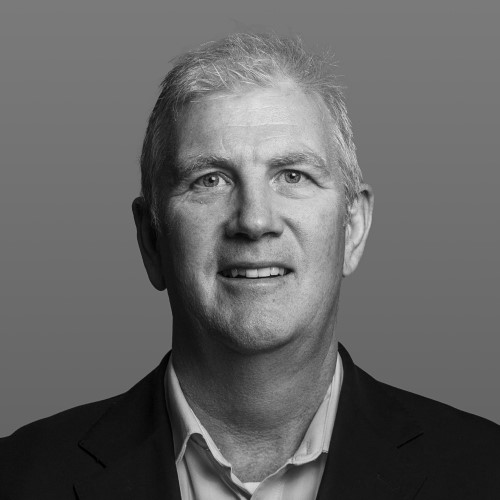 Headshot of a middle-aged man with light skin and short gray hair, smiling confidently while wearing a dark blazer over a collared shirt.
