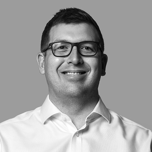Smiling man with short dark hair and glasses, wearing a white dress shirt. The individual appears to be of Caucasian descent.