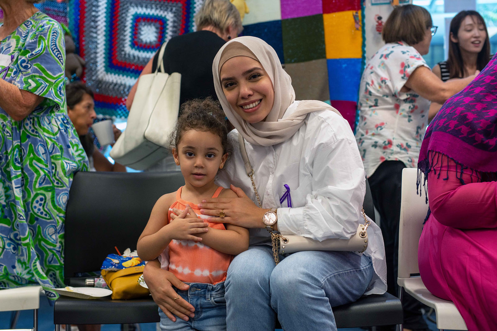A woman wearing a white hijab and a light-colored shirt smiles while sitting next to a young girl in an orange tank top. The girl has curly hair and a joyful expression. They are in a colorful setting with various textile patterns in the background.