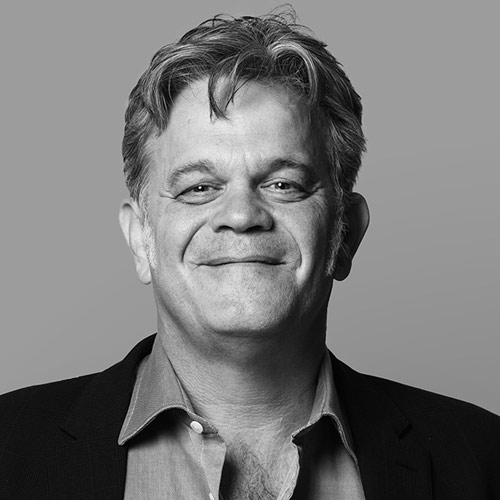 Smiling middle-aged man with light brown hair wearing a dark blazer over a light blue shirt.