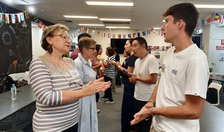 Group of diverse individuals engaged in conversation in a classroom setting. Other participants are visible in the background, all involved in discussions.