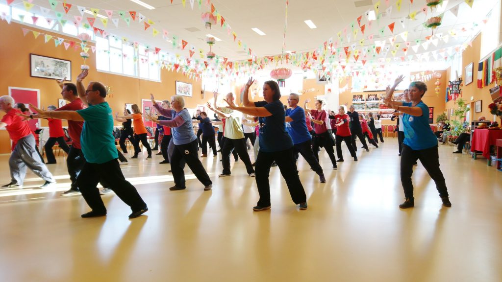 Group of diverse participants engaged in a taichi class, wearing colourful athletic clothing. The participants are focussed as they follow the instructor's movements. Bright decorations hang from the ceiling.