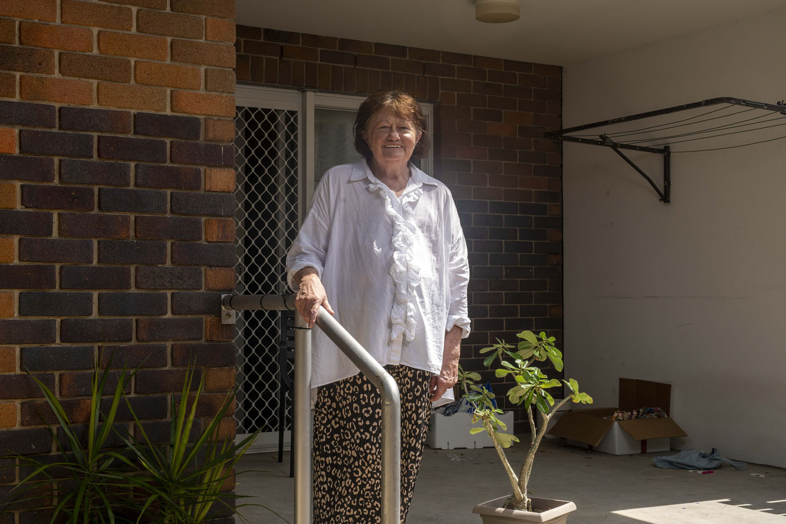Elderly woman with light brown hair standing on a porch, wearing a white button-up shirt and patterned brown pants. She looks directly at the camera smiling, holding onto a metal railing.