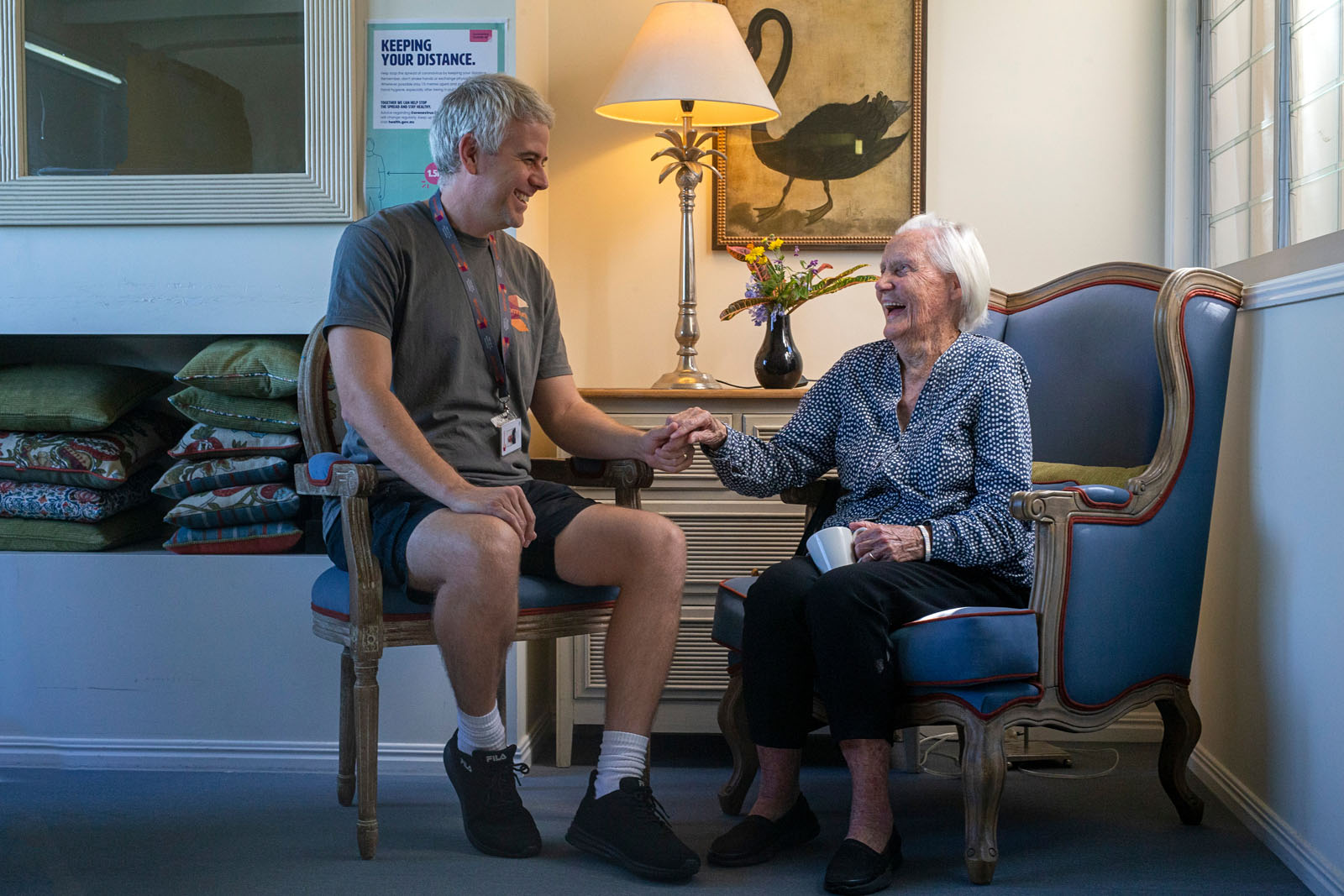 A smiling elderly woman with white hair, wearing a blue patterned blouse and black pants, sits in a chair holding hands with a man. The man, wearing a gray t-shirt and black shorts, has short gray hair and a friendly expression. They are engaged in a warm interaction in a cozy indoor setting.