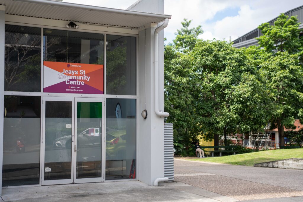 Exterior view of a community center featuring a colorful banner. The entrance has large glass doors and is flanked by greenery. The building is modern and well-maintained, emphasizing community engagement.