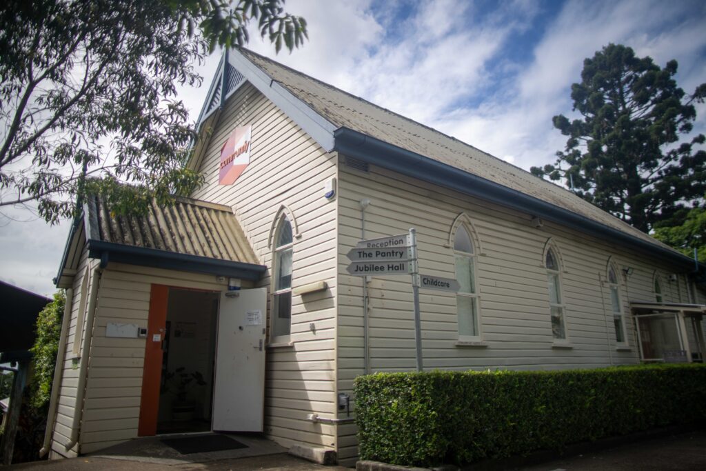 Bardon Community Hub - a white wooden building featuring a peaked roof and arched windows. Green shrubs and trees are visible in the foreground.