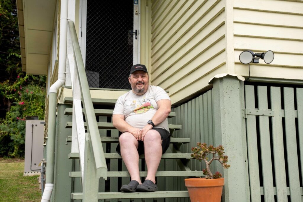 A man with a beard and glasses sits on green stairs outside a house. He wears a light gray t-shirt and black shorts, smiling. A potted plant is beside him.