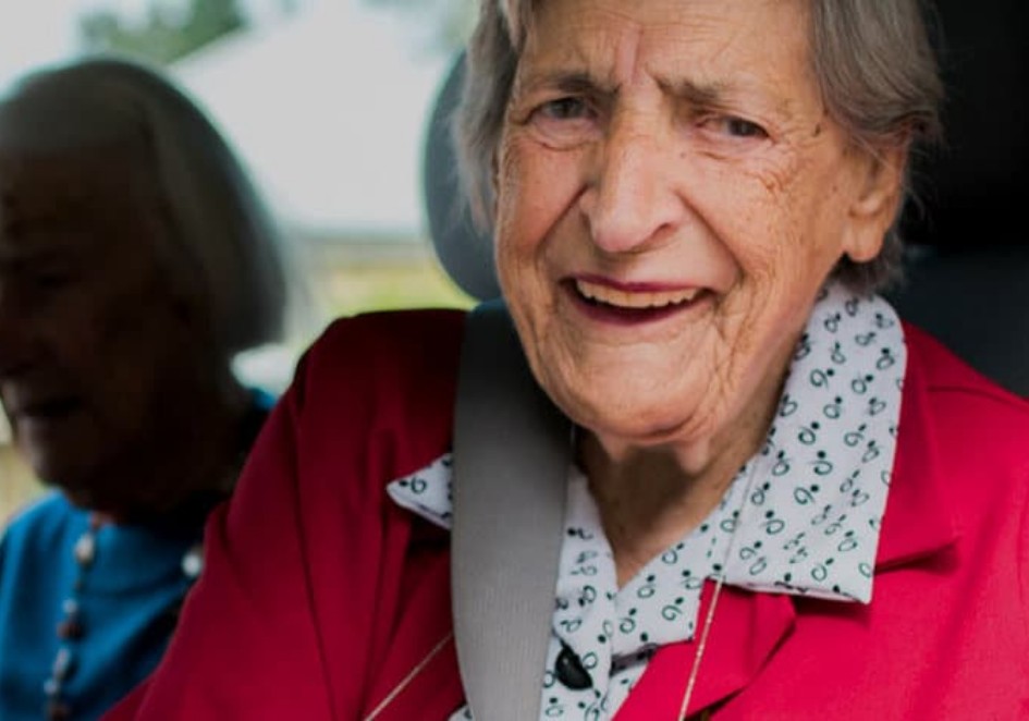 Smiling elderly woman with gray hair wearing a red jacket over a white blouse with a floral pattern. She appears cheerful and is seated in a vehicle, with another person partially visible in the background.