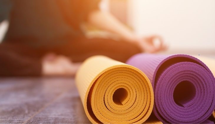 Close-up of a rolled yellow yoga mat and a rolled purple yoga mat on a wooden floor, with a person in the background sitting cross-legged in a meditative pose. The individual is wearing dark clothing and appears focused and relaxed.